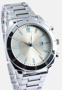 Ασημένιο μεταλλικό ρολόι χειρός με υφασμένο ανοιχτό ασημί καντράν, μπλε δείκτες ωρών και γυαλιστερό λουράκι. Διαθέτει παράθυρο ημερομηνίας και κορώνα.