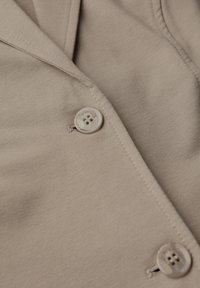 Beige skjorta med knappar som har en slät tygtextur, slagkrage och en lätt, subtil lyster. Knappen har "StreetOne" ingraverat på sig.