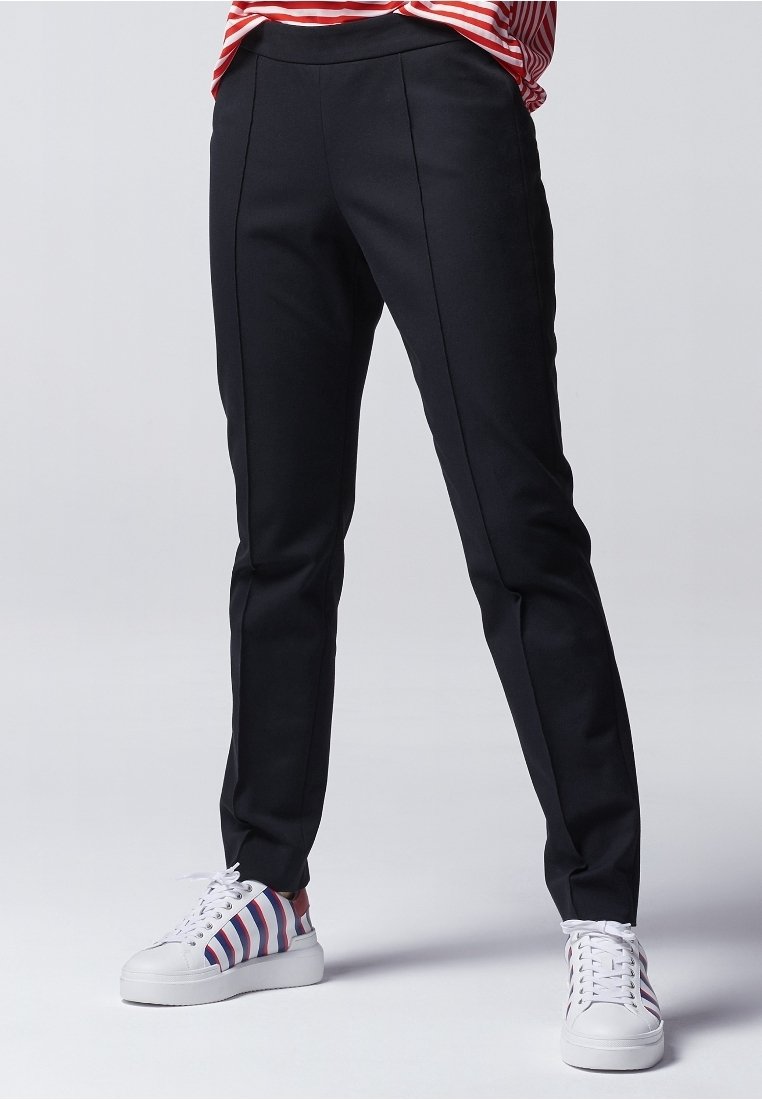Pantalon noir sur mesure avec une texture lisse, présentant des plis à l'avant et une coupe ajustée. Associé à des baskets blanches ornées de rayures rouges et bleues.