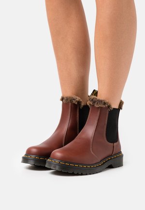 Botas de cuero marrón hasta el tobillo con cuello de piel sintética, paneles laterales elásticos, suela de goma negra y costuras amarillas en contraste.