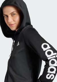 Czarny polar z zamkiem błyskawicznym i podszewką w kapturze. Zawiera białe logo "adidas" na prawej rękawie. Gładki materiał z dopasowanym krojem.