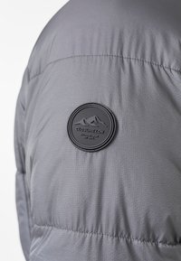 Chaqueta aislante gris con un tejido texturizado, diseño acolchado y una etiqueta circular negra con gráficos de montañas y el nombre de la marca.