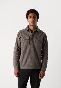 Camisa de botones gris hecha de algodón, con dos bolsillos en el pecho y mangas largas, exhibida en un modelo frente a un fondo simple.