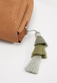 Keltainen mokkasilmälaukku, jossa on ommeltu yksityiskohta ja kolmikerroksinen tupsu vihreänä ja harmaana, kiinnitetty kantolenkkille. Kultaista koriste-nuppia.