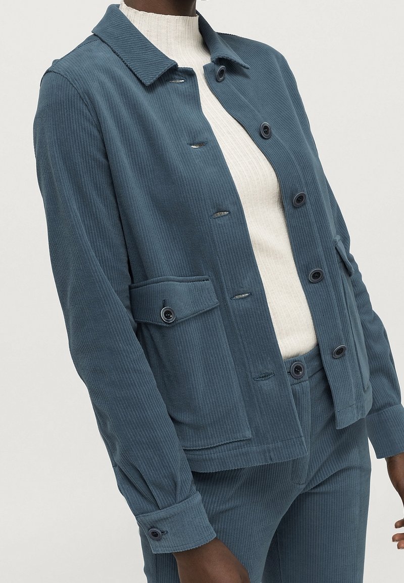 Modrá kordurová bunda s límcem, předními knoflíky, náprsní kapsou a knoflíkovými manžetami, zkombinovaná se světlým žebrovaným topem.