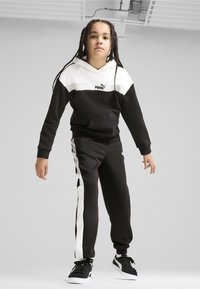 Sudadera en blanco y negro con un bolsillo tipo canguro, combinada con pantalones de chándal negros que cuentan con rayas blancas en los laterales. El material parece suave y cómodo.