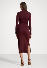 Abito lungo bordeaux a maniche lunghe con colletto alto, vestibilità aderente e spacco laterale, realizzato in tessuto morbido. Indossato con sandali con tacco alto color beige.