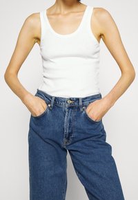 Top blanco sin mangas con escote redondeado, combinado con jeans de mezclilla azul de talle alto. Los jeans cuentan con bolsillos y cierre de botón.