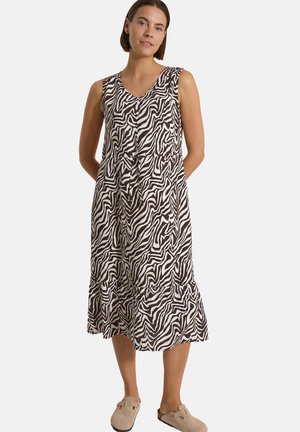 Kvinde iført en ærmeløs knælængde kjole med sort og hvid zebra print og beige slip-on sko, stående med hænderne bag ryggen.