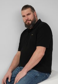 Zwarte poloshirt met een kraag, voorzien van een klein gouden logo op de borst, gecombineerd met blauwe spijkerbroeken. Glad materiaal, casual pasvorm.