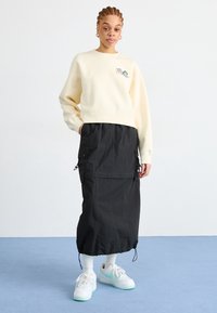 Ljust beige sweatshirt med frontbroderi, kombinerad med en svart, midjelång kjol med cargo-fickor och justerbara snörr. Vita och mintgröna sneakers.