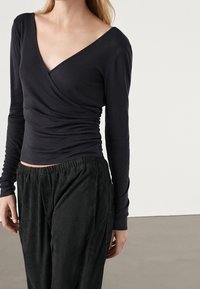 Czarna bluzka z długim rękawem, z głębokim dekoltem w kształcie litery V i marszczonymi detalami po bokach, w połączeniu z czarnymi, luźnymi, teksturowanymi spodniami z elastycznym pasem.