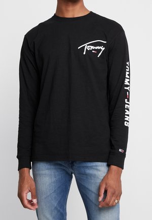 Mężczyzna ubrany w czarną długorękawową koszulkę Tommy Jeans z białym logo na piersi i rękawie, zestawioną z jasnoniebieskimi dżinsami.