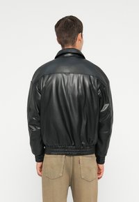 Černá kožená bomber bunda s lesklým povrchem, disponující ribbed lemem a límcem. Záda ukazují uvolněný střih a žádné ozdoby.