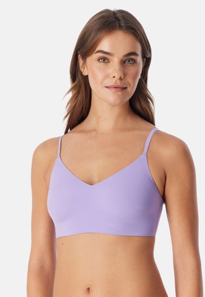 Jonge vrouw met lang bruin haar die een lichtpaarse mouwloze crop top draagt tegen een effen witte achtergrond.