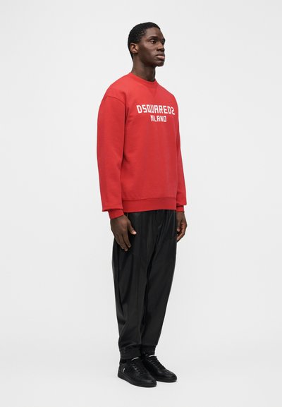 Sudadera roja con texto blanco "DSQUARED2 MILANO", combinada con pantalones deportivos negros y zapatillas negras. Diseño sencillo, corte relajado.