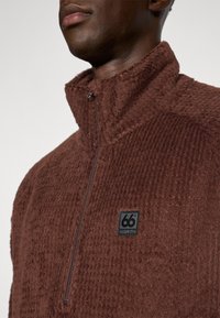 Brauner Fleece-Pullover mit hohem Kragen, strukturiertem Oberflächen und einem Viertelreißverschluss. Mit einem schwarzen Logopatch auf der linken Brust.