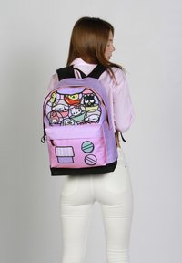 Mochila con un cuerpo lavanda, detalles en rosa y patrones de dibujos animados coloridos en la parte frontal. Tiene un fondo negro y un bolsillo con cremallera.