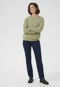 Felpa verde chiaro con una toppa a forma di cuore rosa, abbinata a jeans blu scuro e mocassini neri. Stile casual, taglio rilassato e tessuto morbido.