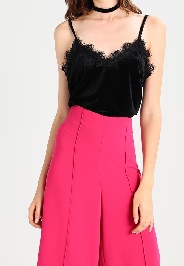 Femme portant une camisole noire ornée de dentelle et un pantalon large taille haute rose vif, avec un collier ras du cou noir.
