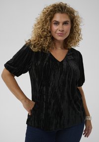 Donna dai capelli ricci che indossa un top nero a V con texture e jeans scuri, in posa con una mano in tasca e un braccialetto di perle al polso.