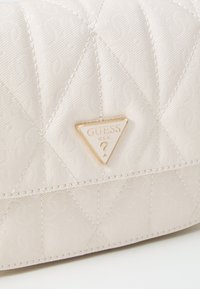 Weiße, gesteppte Handtasche aus strukturiertem Material mit einem goldenen, dreieckigen Logo-Akzent, auf dem der Markenname "GUESS U.S.A." eingeprägt ist.