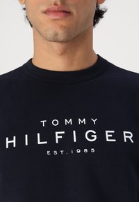 Sweatshirt en coton bleu marine arborant un logo blanc avec l'inscription "TOMMY HILFIGER EST. 1985" en lettres majuscules sur le devant.