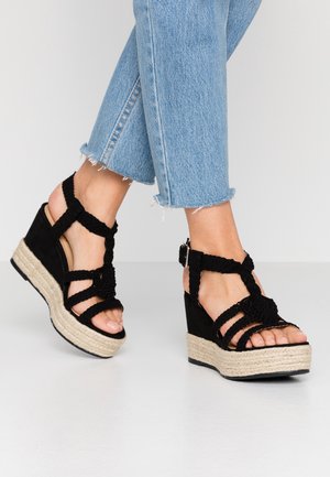 Fødder iført sorte flettede kile-sandaler med jute-såler og ankelremme, kombineret med cropped lyseblå denimjeans.