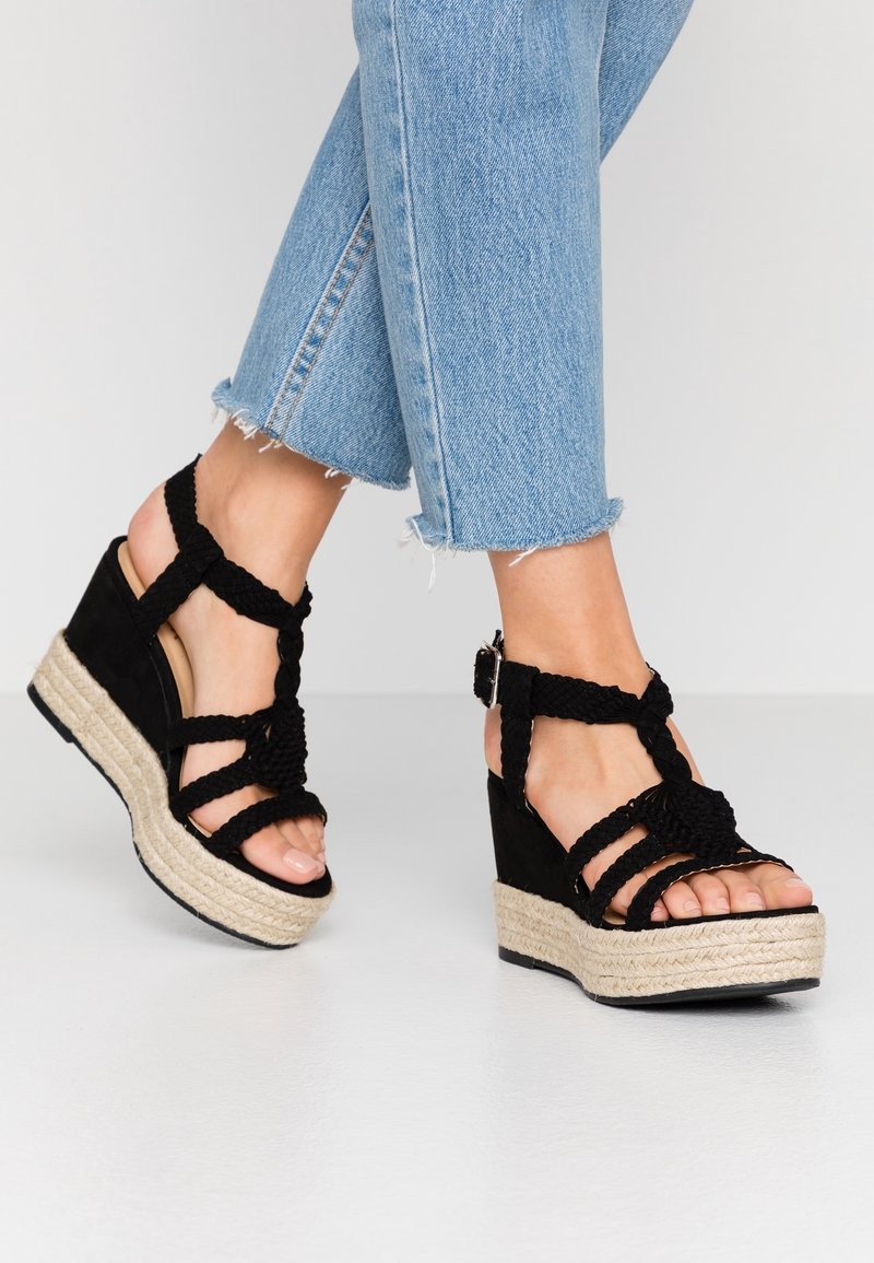 Pieds chaussés de sandales compensées tressées noires avec semelles en corde de jute et brides autour de la cheville, associées à un jean en denim bleu clair raccourci.