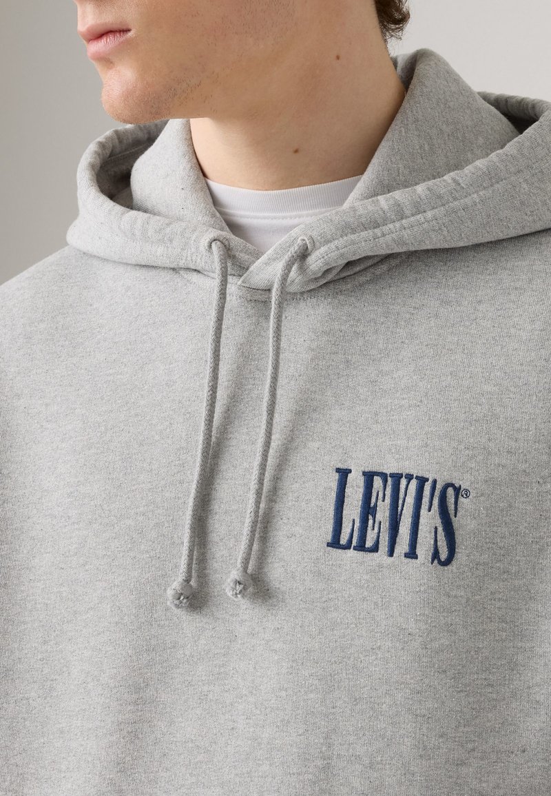 Hoodie en coton gris avec fermeture à cordon, logo brodé bleu marine et texture douce. Design minimalist avec une coupe classique.