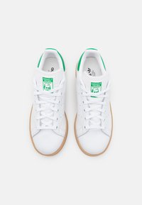 Bílé kožené tenisky se zelenými akcenty a hnědou gumovou podrážkou. Disponují šněrovacím designem a značením na jazyku. Hladký povrch.