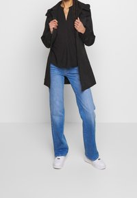 Trench coat leggero nero con chiusura a bottoni e cappuccio, abbinato a jeans ampi blu chiaro e sneakers bianche.