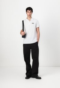 Valkoinen polo-paita lyhyillä hihoilla, musta logo, yhdistettynä mustiin väljälahkeisiin housuihin ja mustiin kenkiin. Malli pitää pientä mustaa laukkua.