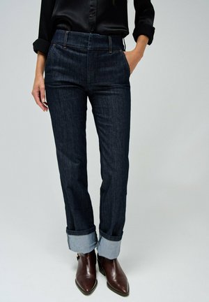 Personne portant un jean en denim foncé retroussé, une chemise noire boutonnée avec manches retroussées, et des bottes en cuir marron à bouts pointus.