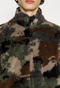 Fleecejakke med camouflage-mønster i grøn, brun og sort. Har høj krave og lynlås-lukning, med en blød, tekstureret overflade.