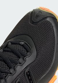 Crne atletske cipele s mrežastim gornjim dijelom, teksturiranom površinom i naglašenim narančastim gumeni potplat. Reflektivni detalj na vezicama.