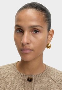 Suéter beige texturizado con cuello redondo y un botón oscuro. Pendiente de aro dorado. Cabello liso recogido hacia atrás, fondo neutro.