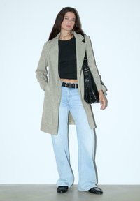 Ženska stoji ob belem zidu, oblečena v bež kariran plašč, črn crop top, svetlo modre jeans hlače, črn pas, črno torbico in črne čevlje.