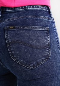 Mörkblå denimjeans med en framträdande bakficka med en böjd söm. Fickan har en liten svart etikett med varumärket "Lee."