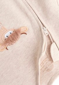 Tutina beige con zip, caratterizzata da un ricamo di una scimmia in peluche e dettagli in pelliccia marrone lunga, realizzata in un tessuto morbido e testurizzato.