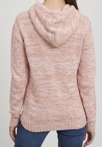 Kvinna som bär en rosa och vit melerad stickad huvtröja med långa ärmar, stående med ryggen mot kameran mot en neutral bakgrund.