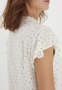 Weiße Bluse mit kurzen, gerafften Ärmeln, versehen mit einem Blumenmuster in Gelb und Pink. Leichter Stoff mit strukturierter Oberfläche.
