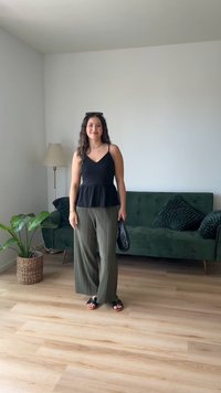 Svart peplum topp, grønne, plisserte vidben bukser, svarte sandaler, holder en strukturert svart veske, står i et lyst rom med en grønn sofa.