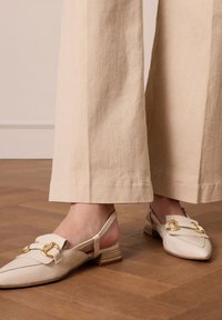 Witte leren slingback schoenen met twee decoratieve gespen, een puntige neus en een lage gestapelde houten hak, gecombineerd met lichtbeige wijde broekspijpen.