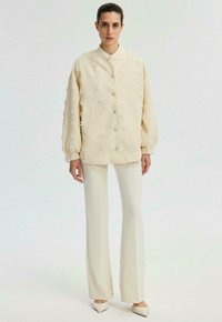 Veste couleur crème avec motifs floraux texturés, devant boutonné et poignets élastiques, associée à un pantalon crème slim et des chaussures pointues.