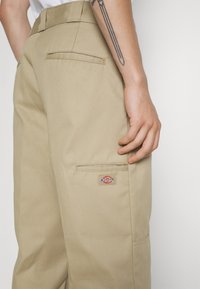 Pantalon de travail beige à coupe droite, avec poche arrière visible et une étiquette de marque sur le côté droit. Fabriqué en tissu résistant avec une texture lisse.