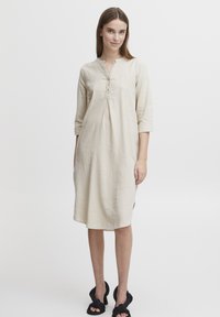 Robe en lin beige clair avec un col henley, des manches trois-quarts, des fentes sur les côtés et des boutons sur le devant. Portée avec des sandales noires.