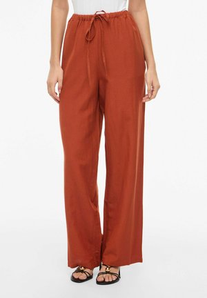 Kobieta w szerokich spodniach o kolorze rdzy z troczkiem w pasie i czarnych, odkrytych sandałach na palce ze złotymi akcentami na białym tle.