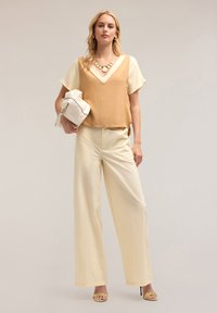 Lichtbeige mouwloze top over een romige blouse, gecombineerd met brede crèmekleurige broek en gele haksandalen. Houdt een witte handtas vast.