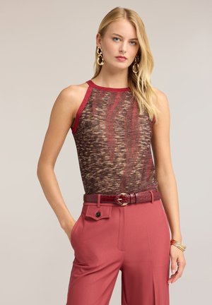 Blonde Frau in ärmellosem braun-rot gemustertem Oberteil, hoch taillierte rote Hose mit Gürtel, goldene Ohrringe und Armbänder, stehend mit einer Hand in der Tasche.